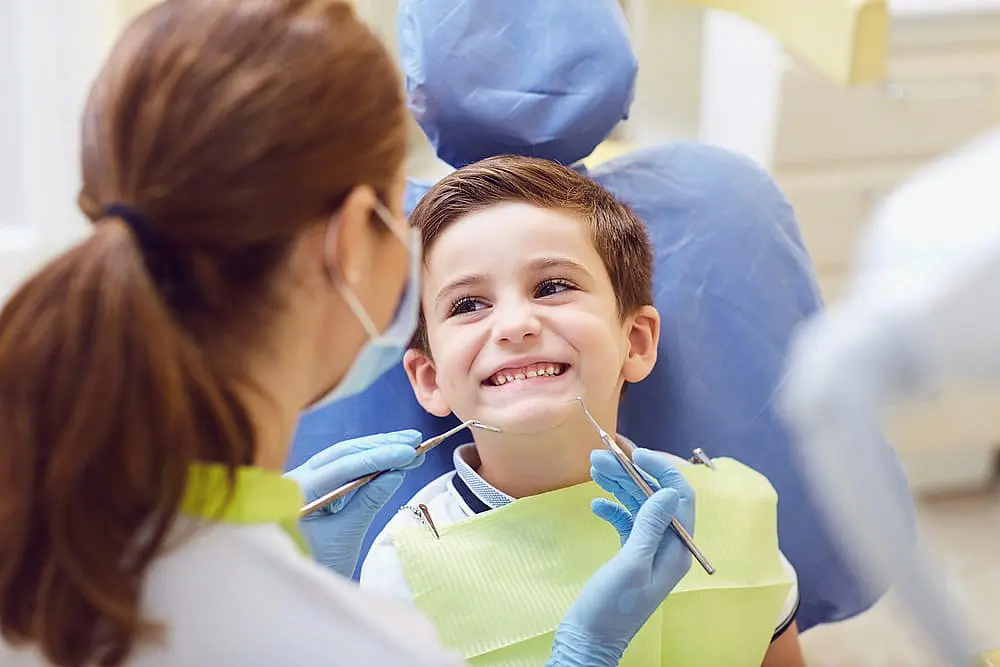 A dentist examines a smiling child’s teeth in a dental office, where orthodontic appliances are neatly arranged nearby. The child is wearing a green bib, while the dentist, holding dental tools, ensures everything is in place for optimal oral health in Leominster, MA