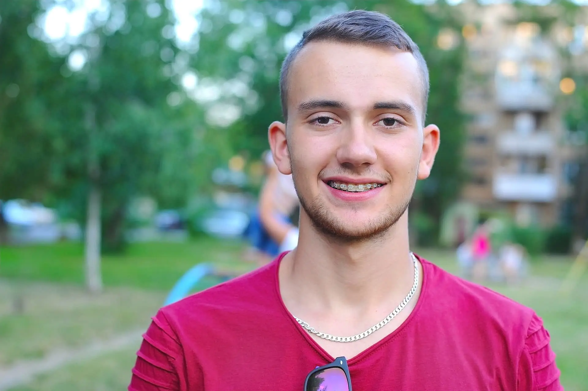 A young man with clear braces and short hair smiles at the camera in a park, showcasing adult orthodontics with confidence. He's wearing a red shirt and silver chain, set against a backdrop of blurred trees and buildings in Leominster and Gardner, MA