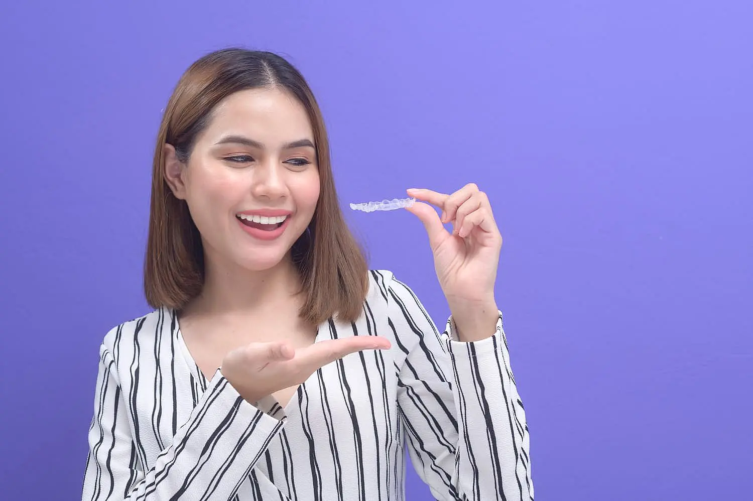 A woman smiles while holding a clear Invisalign dental aligner in one hand, pointing at it with the other, against a purple background in Leominster and Gardner, MA