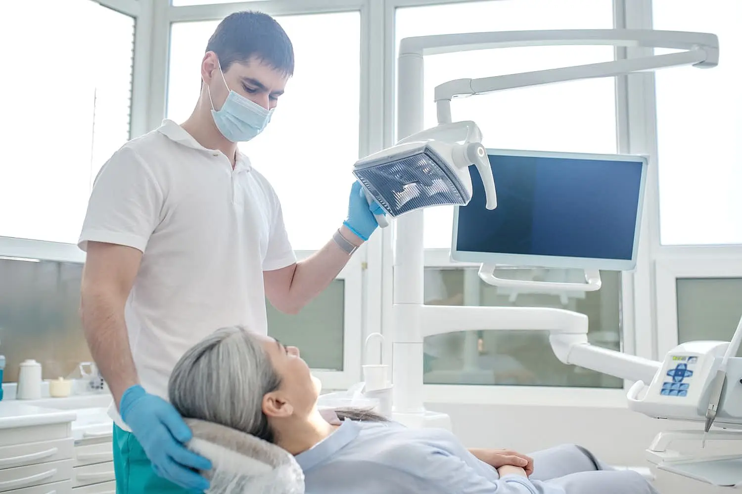 A dentist wearing a mask and gloves adjusts a dental light above a patient in the dental chair, preparing for an adult orthodontics procedure. The room, filled with dental equipment, is brightened by large windows in Leominster or Gardner, MA