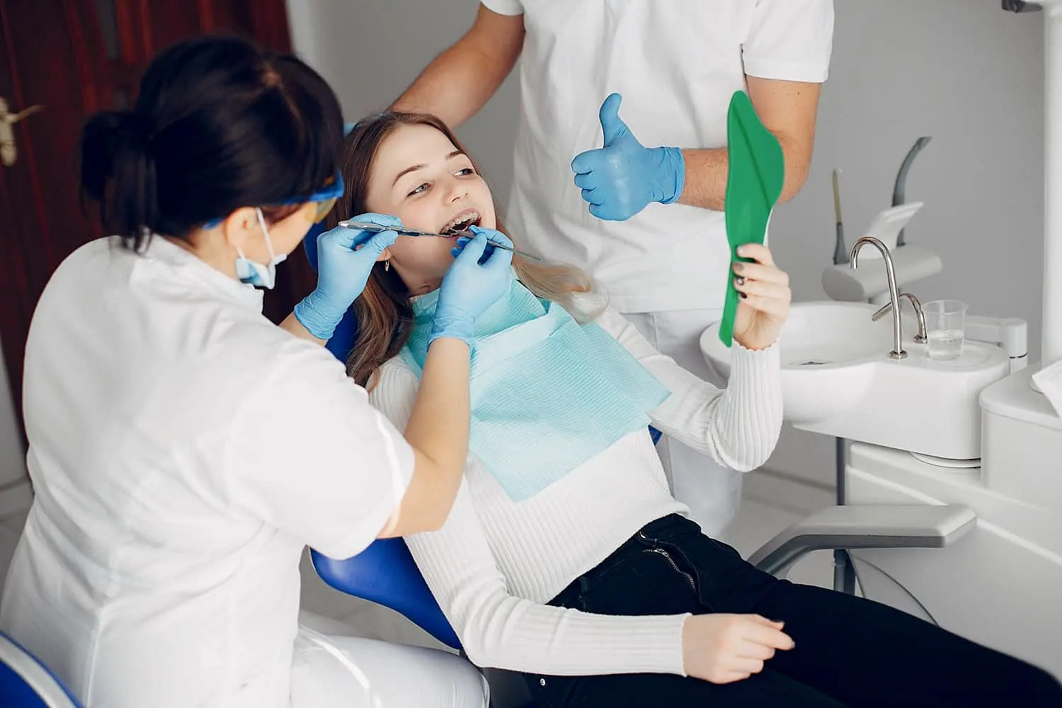 A patient sits in a dental chair holding a mirror while a dentist and an assistant adjust her orthodontic appliances. The assistant wears a mask, and the dentist gives a thumbs-up in Leominster, MA