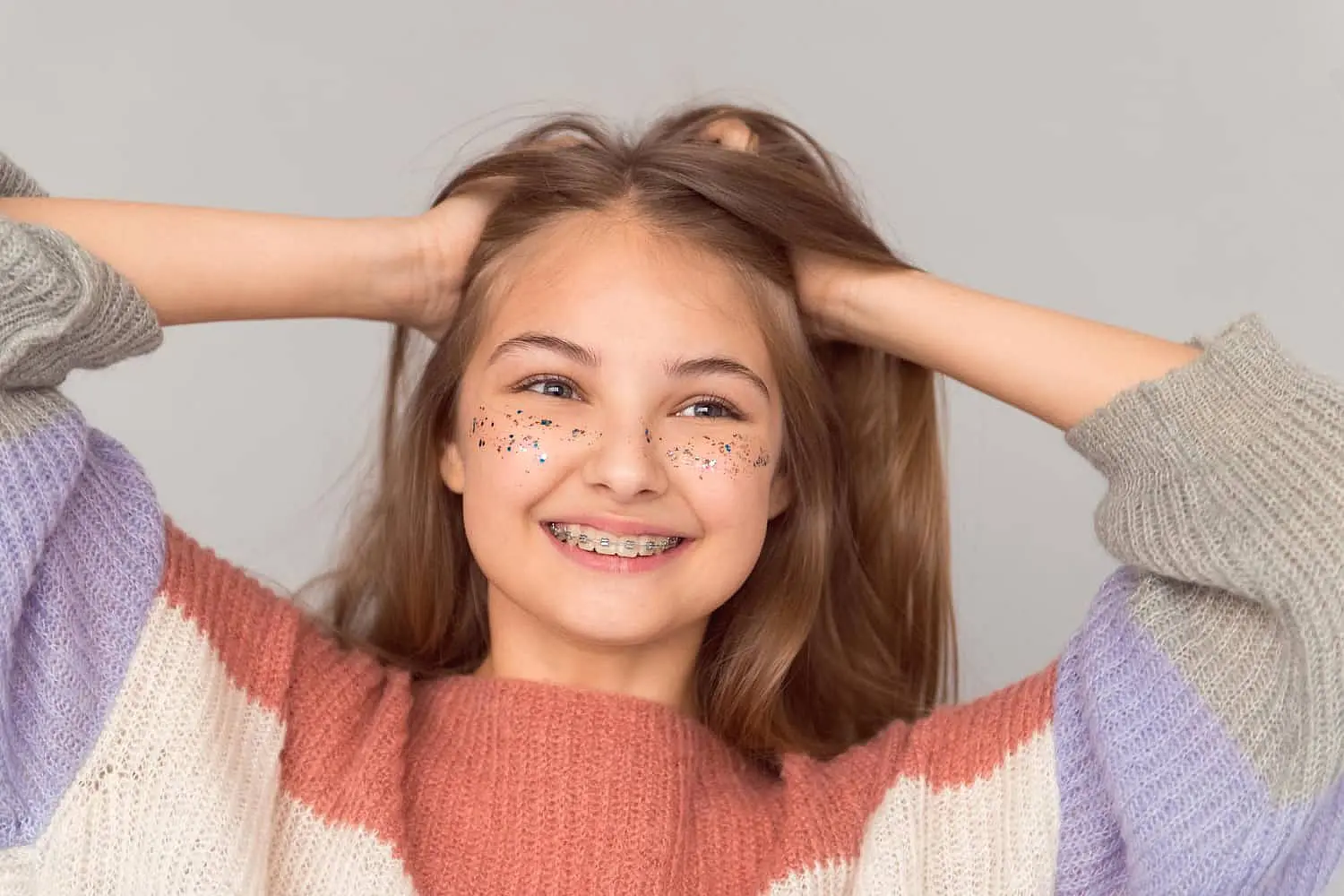 Girl with braces, a testament to her recent visit to the children’s orthodontist, has glitter on her cheeks. She's smiling with hands in hair, wearing a striped sweater against a plain background in Leominster and Gardner, MA