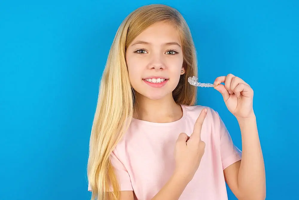 A girl with long blonde hair points to a clear dental aligner against a blue background, embodying the expertise of a children's orthodontist, all while wearing a light pink shirt in Leominster and Gardner, MA