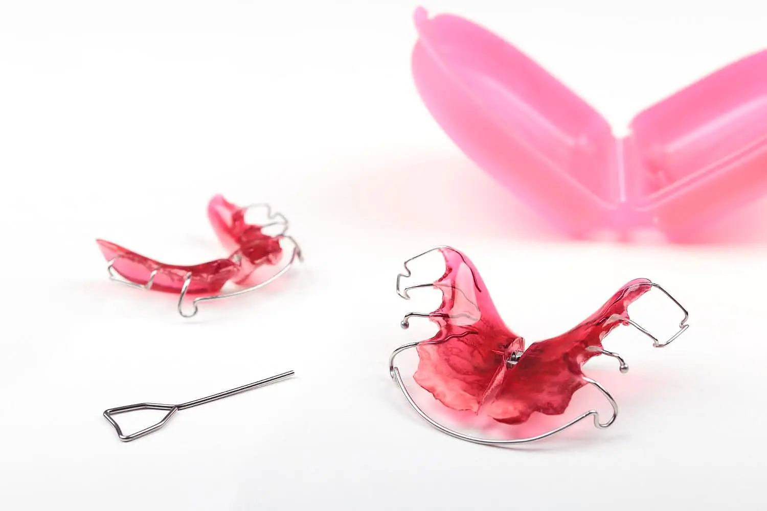 Two red dental retainers with metal wires sit on a white surface, accompanied by a small tool and an open pink case. Perfectly tailored for children’s orthodontist needs, this set combines practicality with a touch of whimsy in Leominster and Gardner, MA