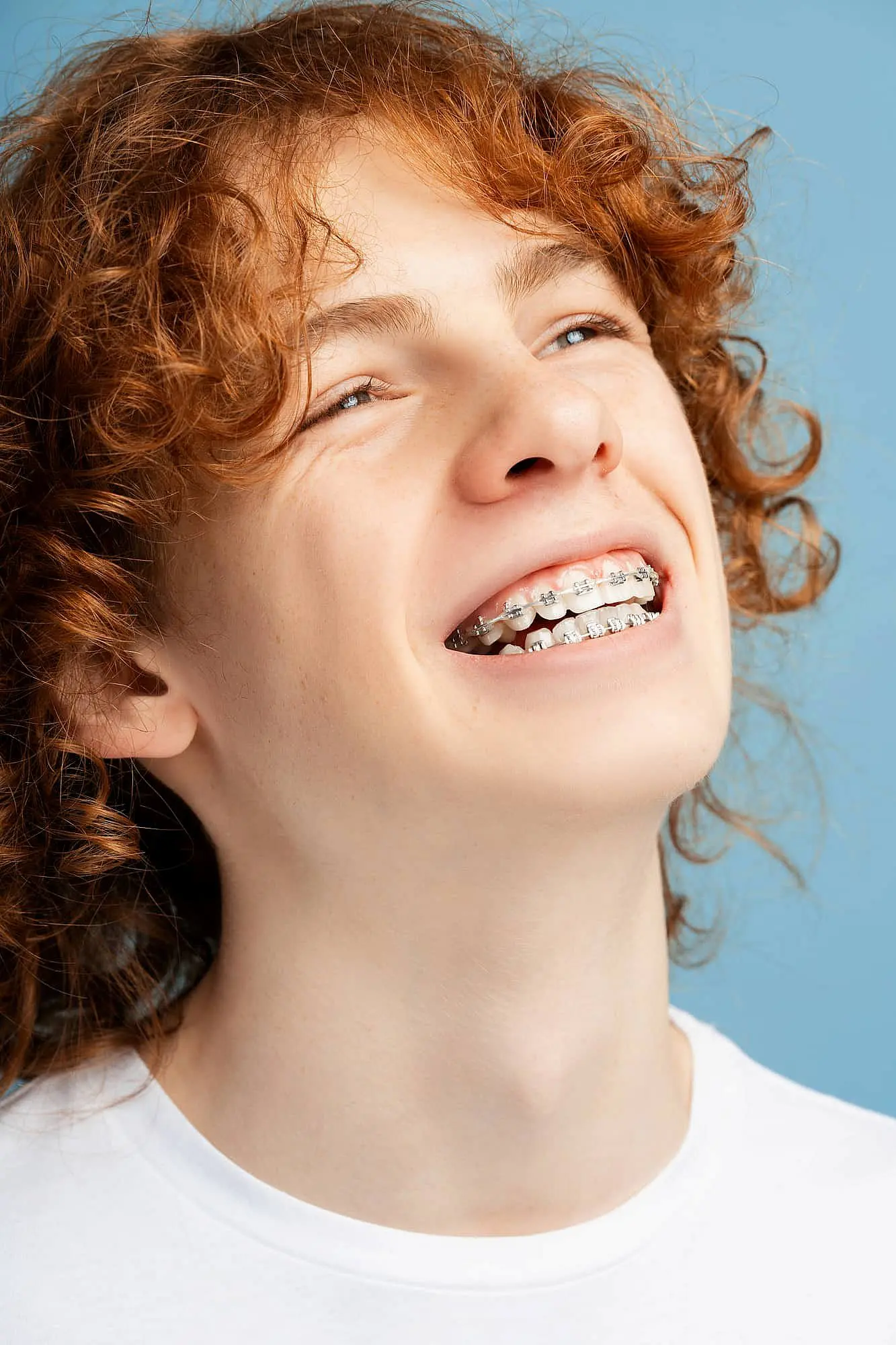 A person with curly red hair is smiling brightly, proudly showcasing their braces as a testament to adult orthodontics, all set against a vibrant blue background in Leominster and Gardner, MA