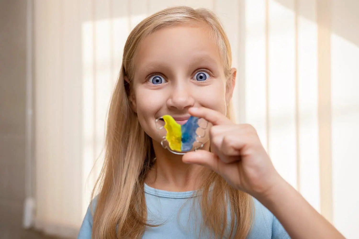 A girl holds a colorful orthodontic appliance, smiling brightly as she showcases her dental retainer in front of her mouth in Leominster, MA