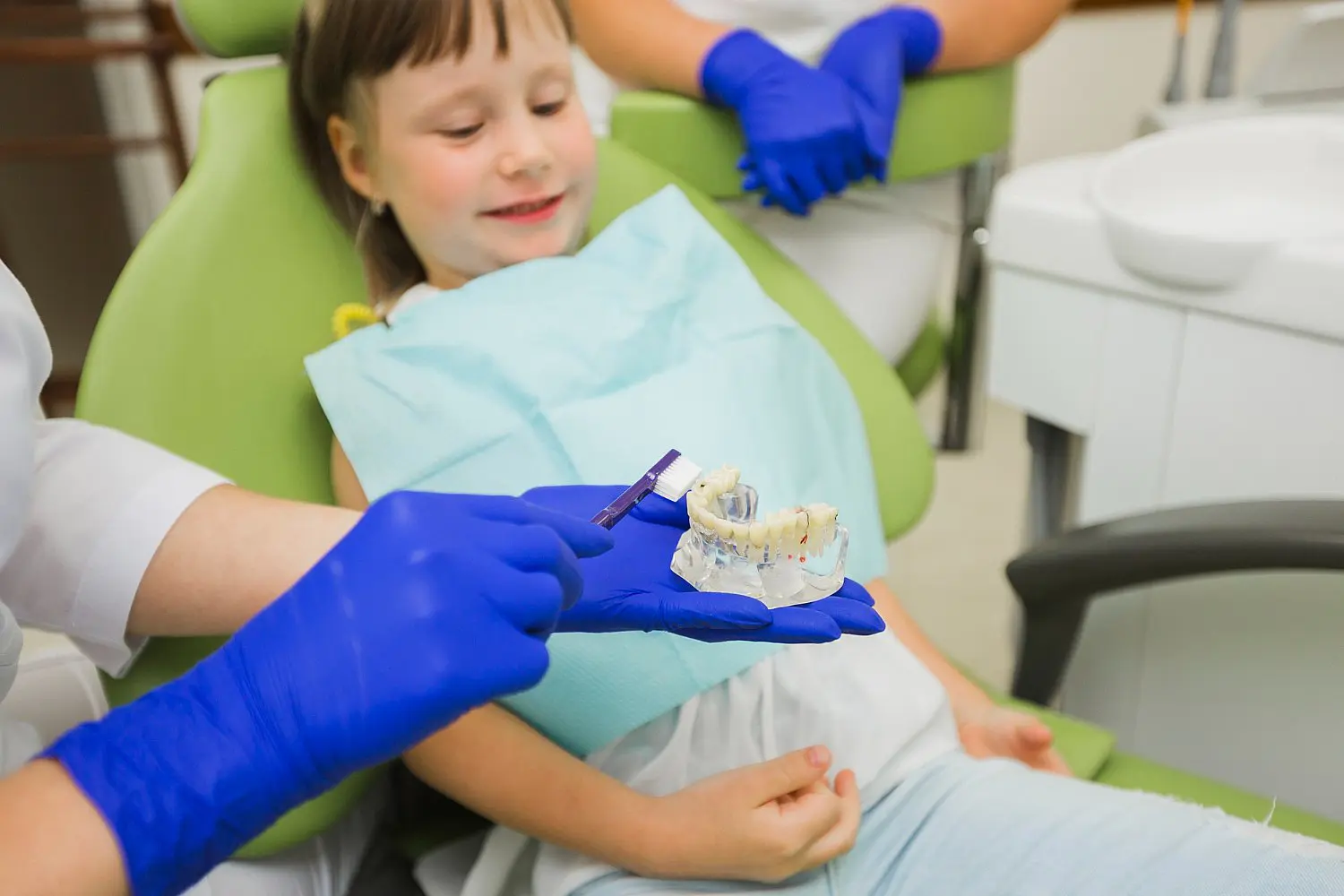 A child in a dental chair keenly observes an orthodontist with blue gloves, holding a dental model and toothbrush to expertly demonstrate children’s oral hygiene techniques in Leominster and Gardner, MA