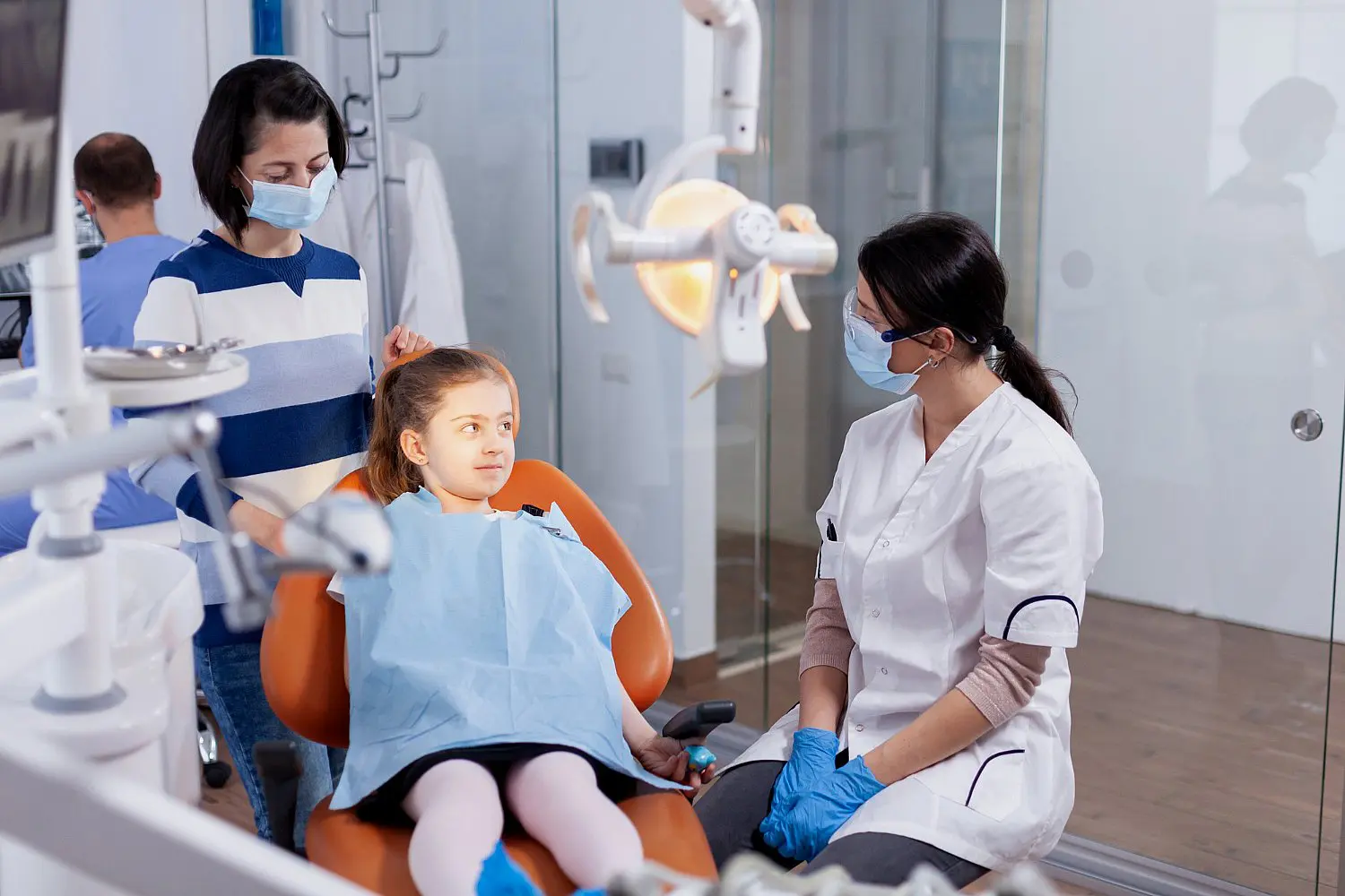 A children's orthodontist speaks to a child sitting on an orange dental chair, accompanied by an adult. Both the orthodontist and the adult are wearing masks in a dental office in Leominster and Gardner, MA