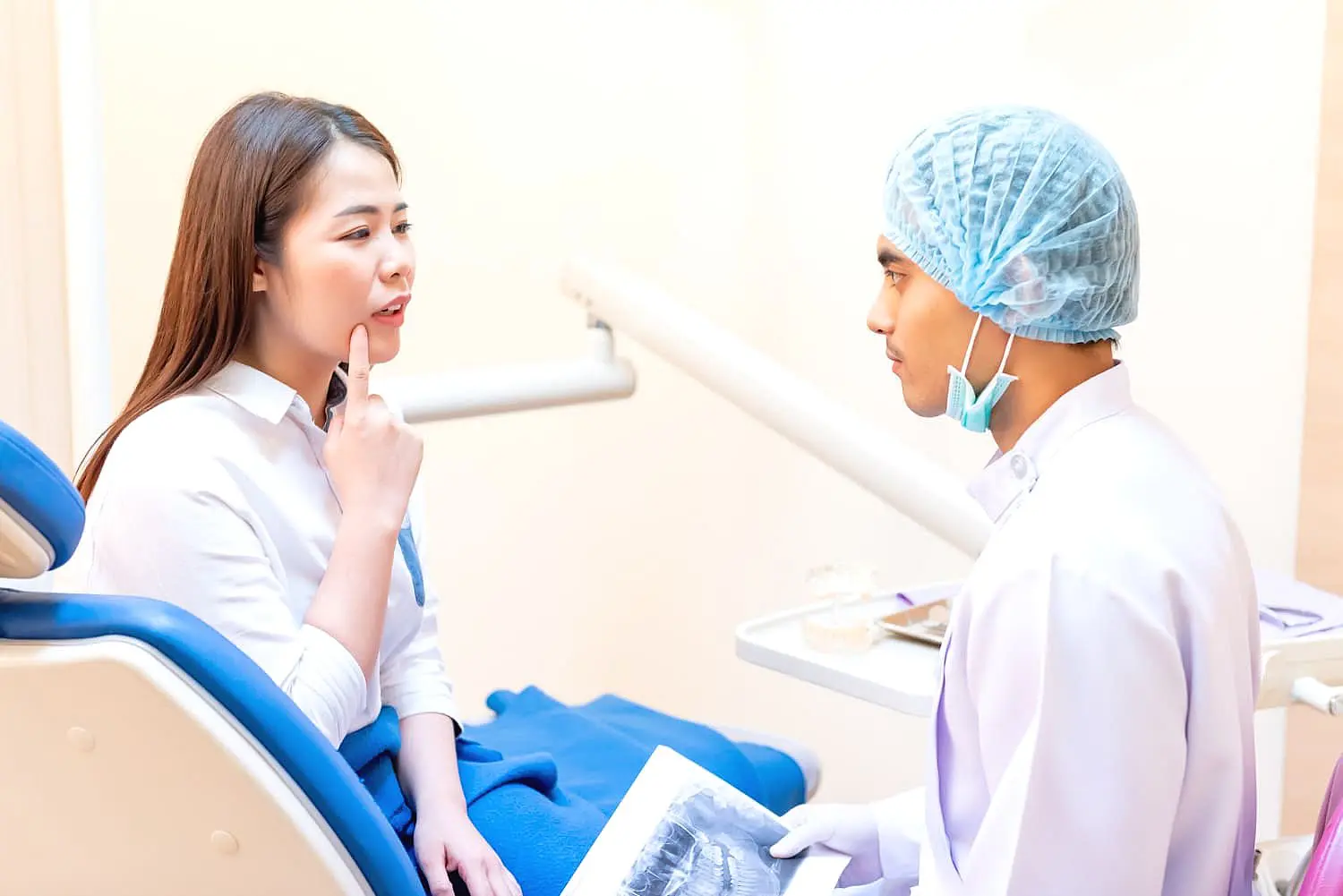 A woman in a dental chair points to her mouth, discussing tongue thrust issues with a dentist who is holding an X-ray. The dentist, donning a hair cap and mask, listens attentively in Leominster and Gardner, MA