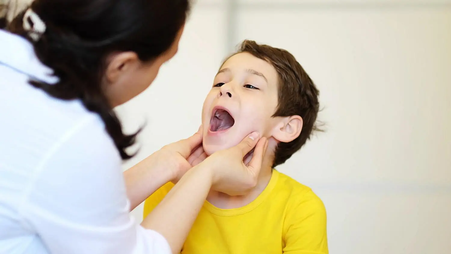 A child in a yellow shirt opens his mouth wide, possibly showing signs of tongue thrust, as a woman gently examines his throat in Leominster and Gardner, MA