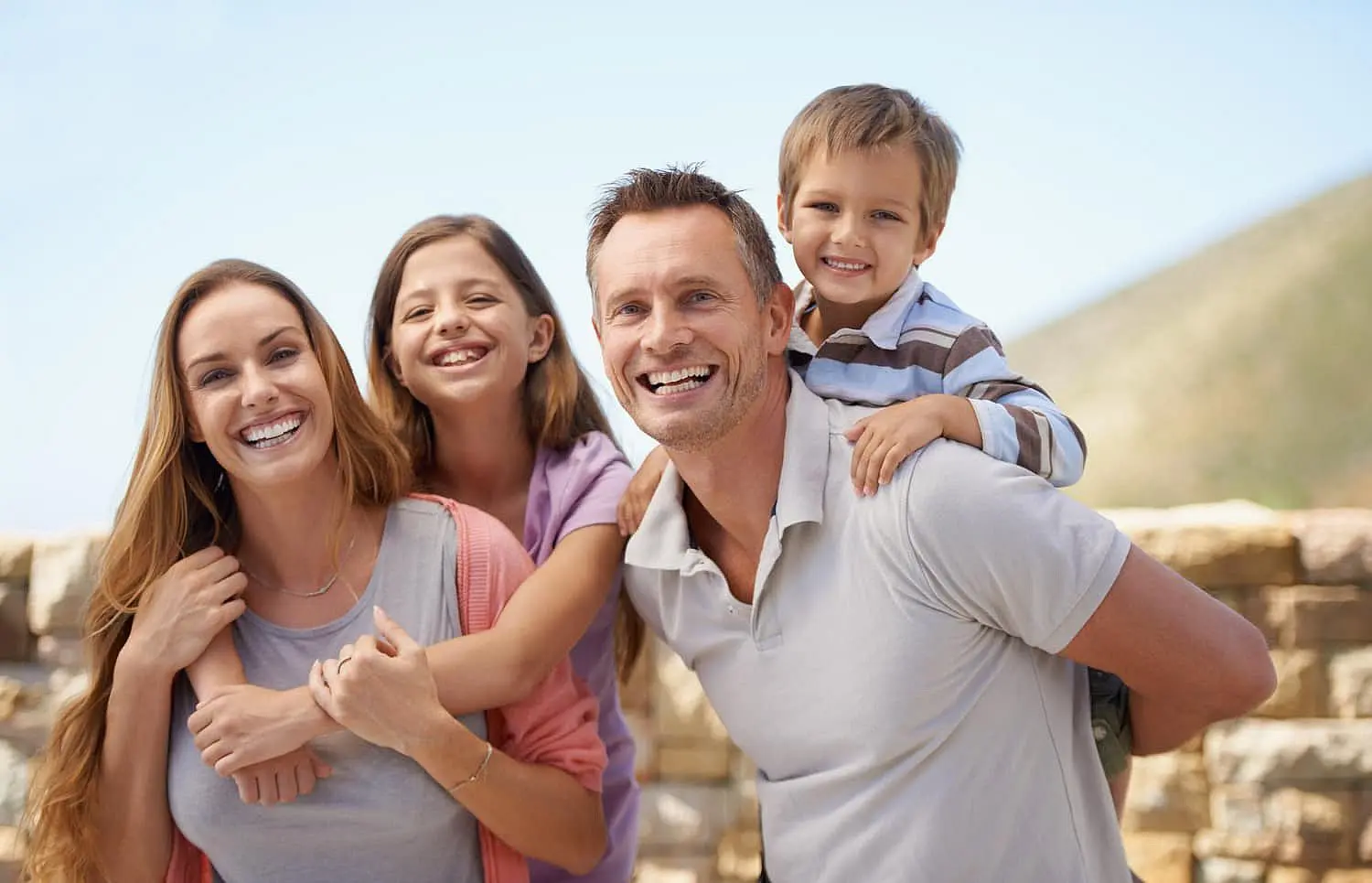 A family of four, with a woman, man, girl, and boy, pose with radiant smiles outdoors against a blurred backdrop of a stone wall and blue sky—a testament to the exceptional care they received at Smile Orthodontics in Leominster or Gardner, MA