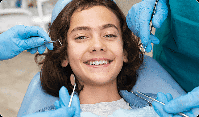 A child lies on a dental chair, smiling brightly, as multiple gloved hands with dental tools gently surround their face. It's a friendly children's orthodontist visit that ensures young smiles grow healthy and straight in Leominster and Gardner, MA