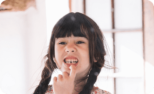 A child with long dark braids smiles broadly, proudly pointing to a gap in her teeth. Standing indoors with soft lighting, she clearly delights in her recent visit to the children's orthodontist in Leominster and Gardner, MA