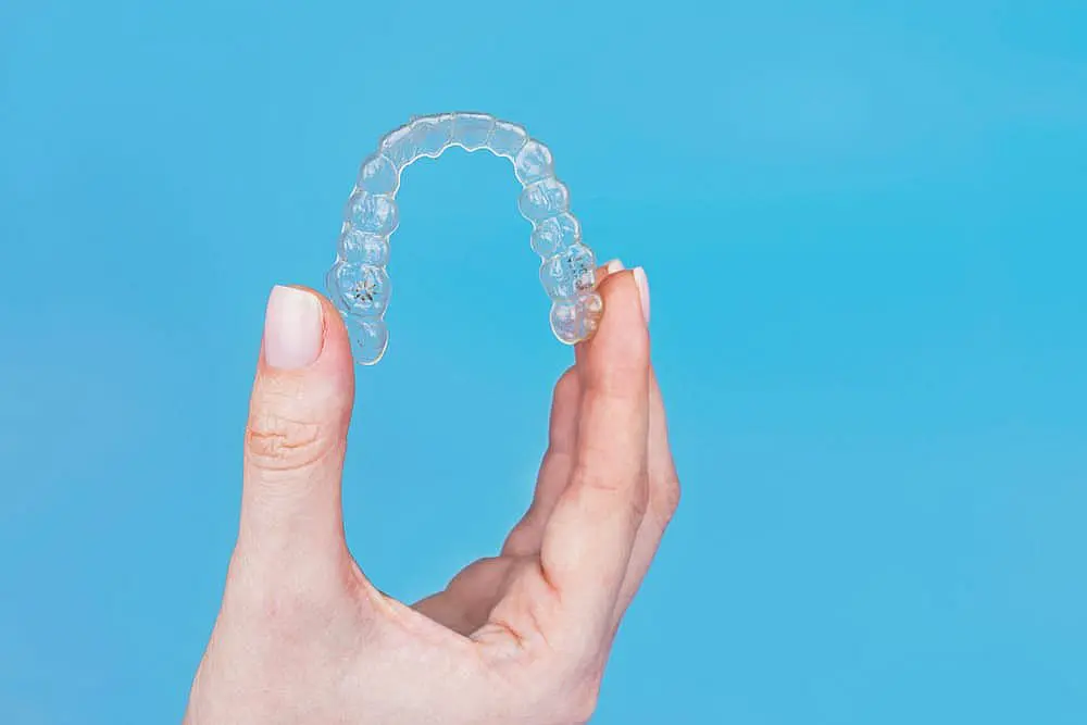 A hand holds a clear dental aligner against a tranquil blue background, subtly hinting at the discreet and effective solution that Invisalign offers. Curious about Invisalign cost? Discover how these aligners can transform smiles with invisible precision in Leominster and Gardner, MA