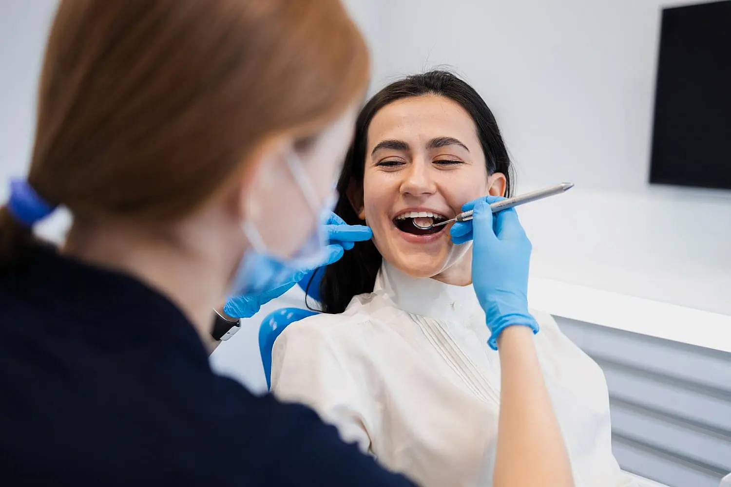 The dentist in blue gloves examines a smiling patient with dental tools, discussing the benefits of adult orthodontics in the clinic in Leominster and Gardner, MA