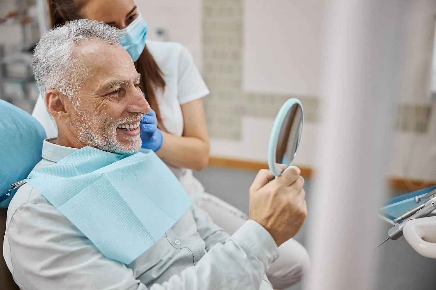 An older man with gray hair, wearing a dental bib, smiles while looking in a hand mirror at the dentist's office. A dental professional in a mask stands beside him, showcasing the results of adult orthodontics in Leominster or Gardner, MA