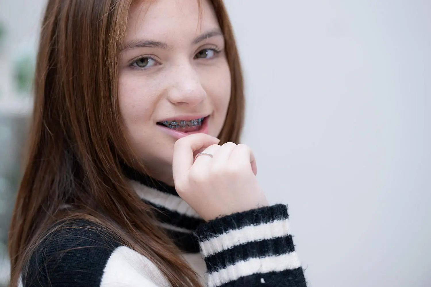 Teen girl with long hair and metal braces smiles while lightly biting her fingernail, dressed in a black and white striped sweater against a plain background in Leominster, MA