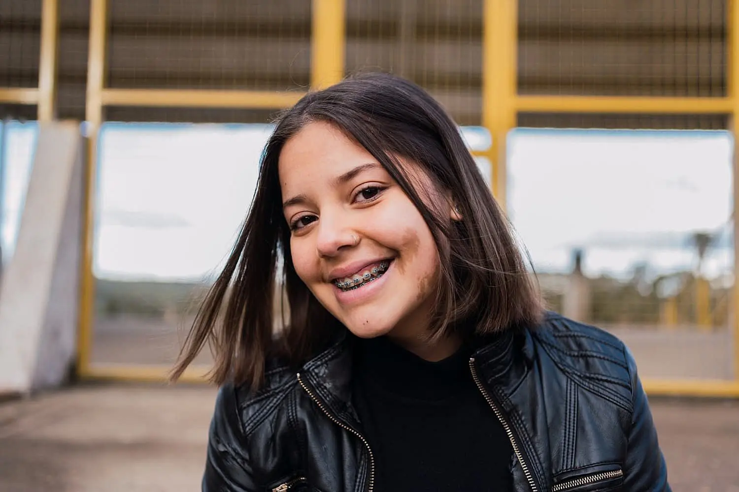 A cheerful girl with braces, wearing a sleek black leather jacket, stands outdoors against a vibrant yellow structure in Leominster and Gardner, MA