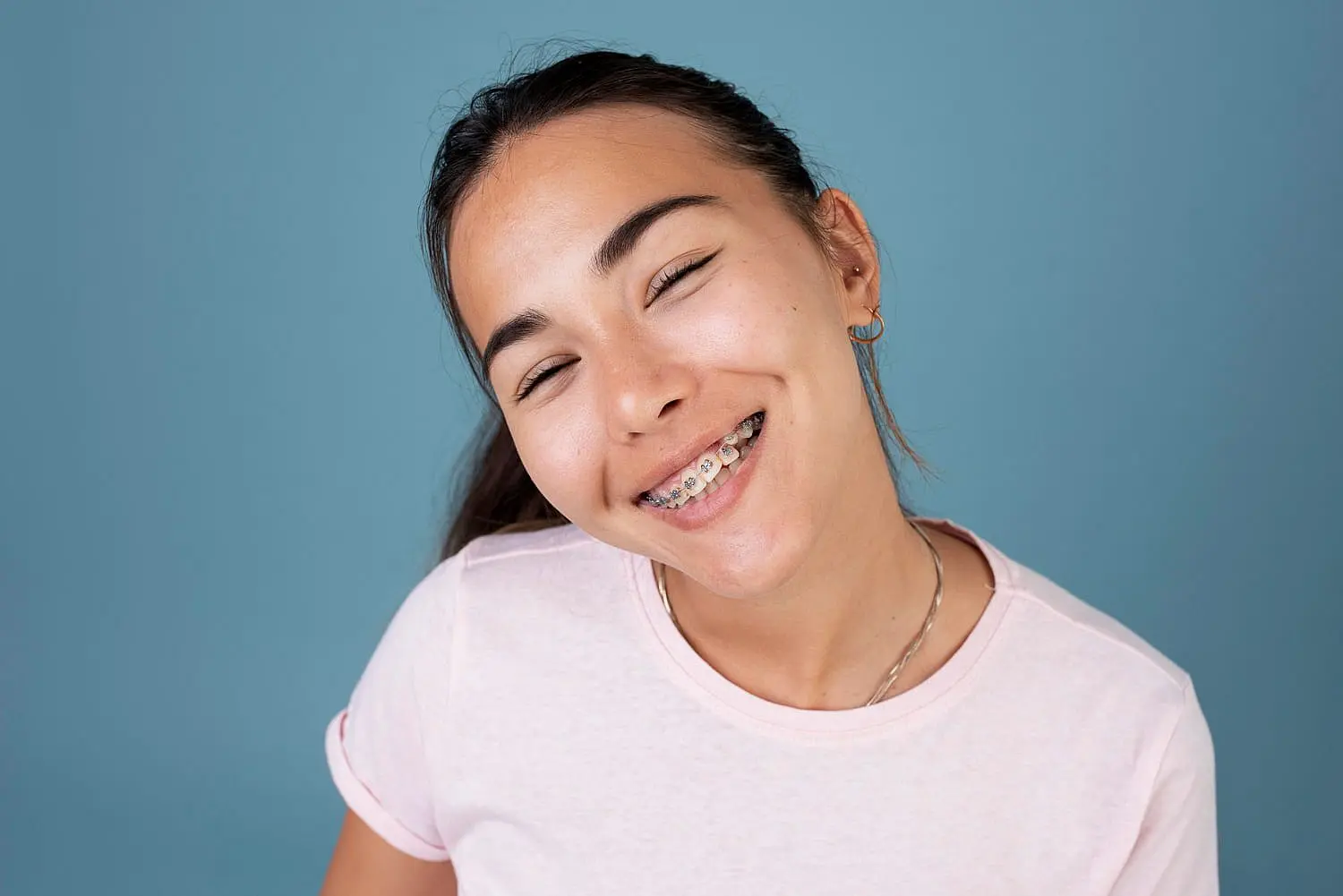 A young person beams with confidence, showcasing their braces while wearing a pink shirt against a light blue background in Leominster and Gardner, MA