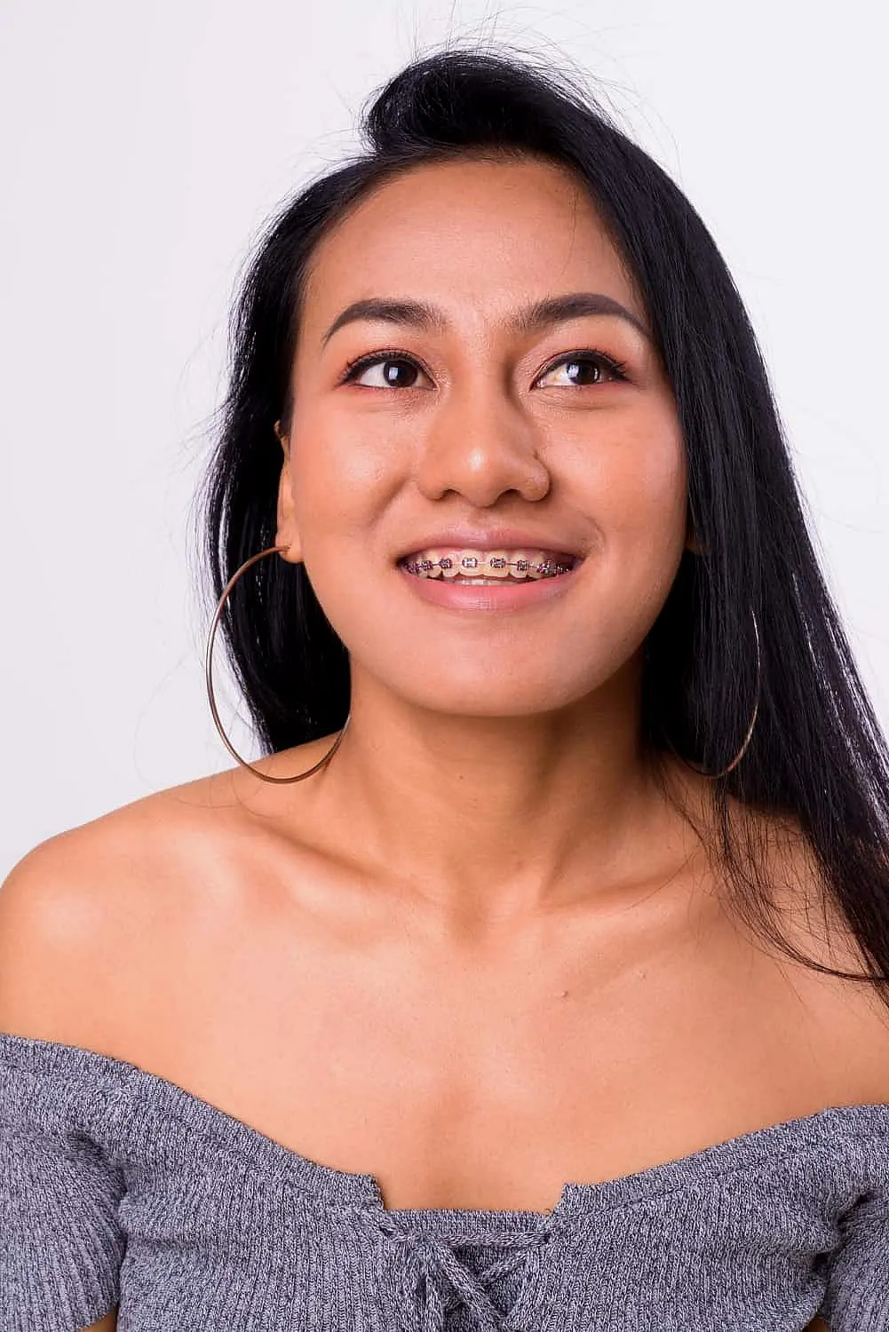 A woman with long hair and metal braces is wearing hoop earrings and a gray off-the-shoulder top, looking slightly upward against a plain white background in Leominster or Gardner, MA