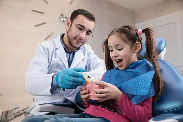 A children’s orthodontist in a white coat and gloves shows a dental model to a smiling girl seated in a dental chair, wearing a blue bib in Leominster and Gardner, MA