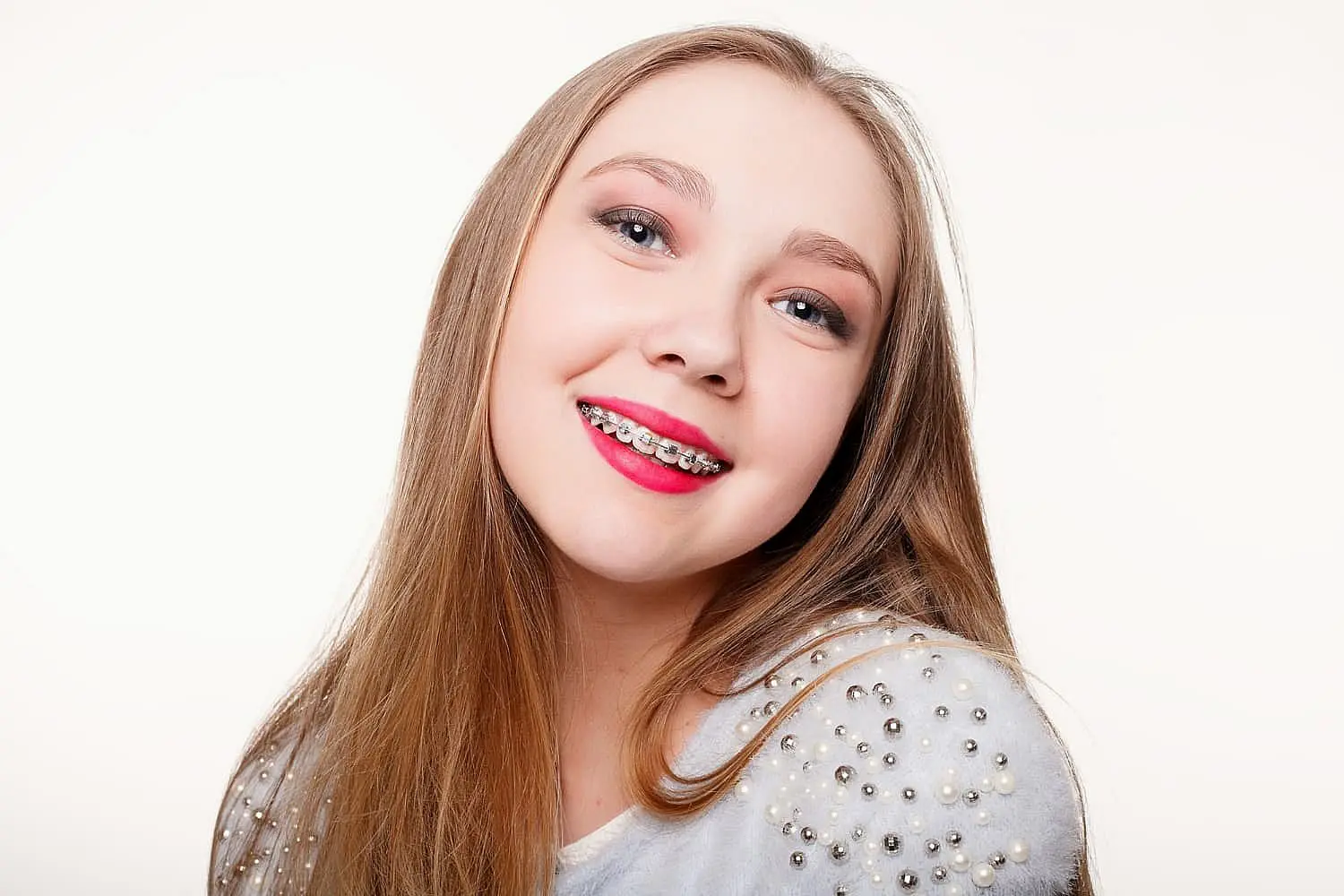A smiling young person with braces, showcasing the brilliance of adult orthodontics, stands against a plain white background. Their long hair frames a pearl-studded top, adding elegance to their look in Leominster and Gardner, MA