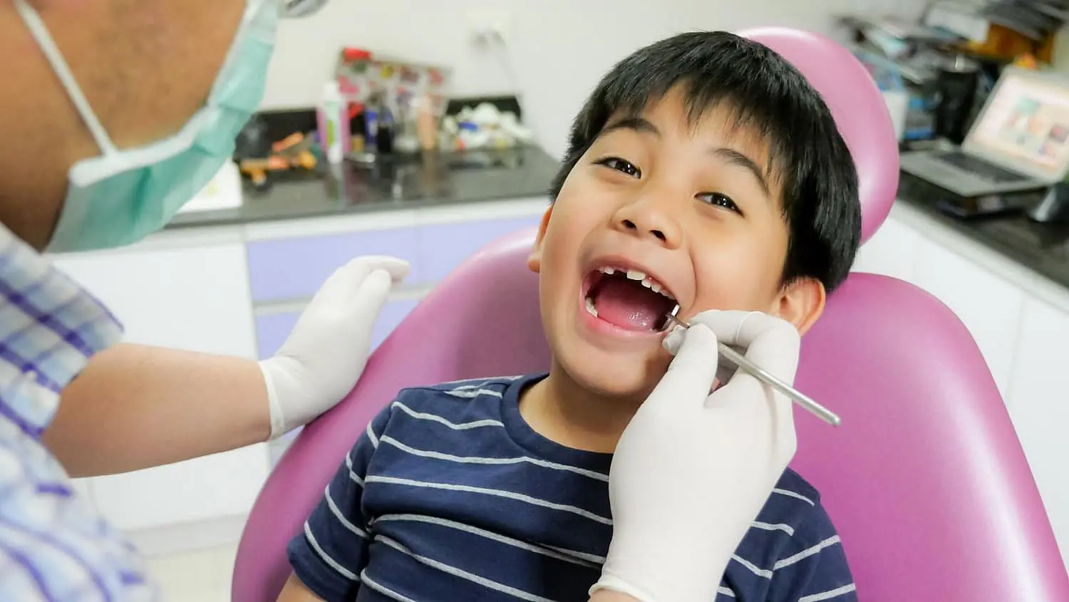 A child sits happily in a dentist's chair, grinning with their mouth open as the children's orthodontist, wearing gloves and a mask, carefully examines their teeth in Leominster and Gardner, MA