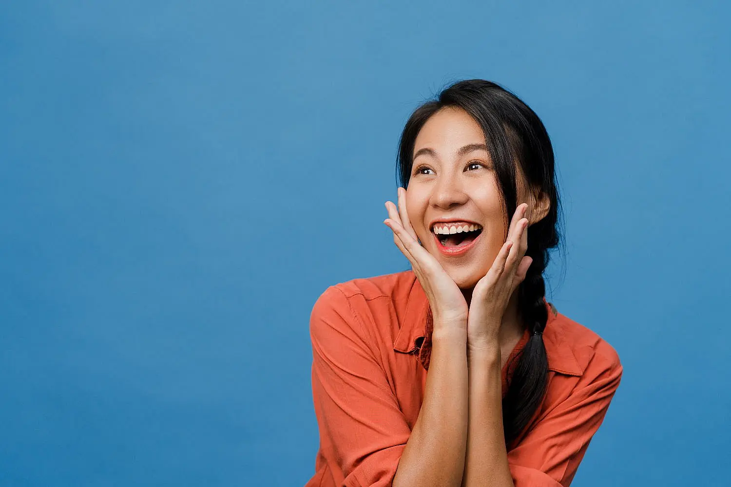 A person in an orange shirt is smiling with hands on cheeks against a solid blue backdrop, radiating joy as if illuminated by LightForce in Leominster or Gardner, MA