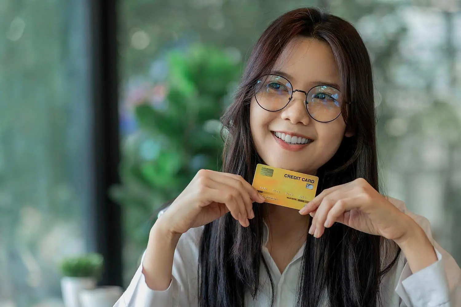 A person with long hair and glasses smiles while holding a credit card, possibly ready to explore a braces payment plan in Leominster and Gardner, MA