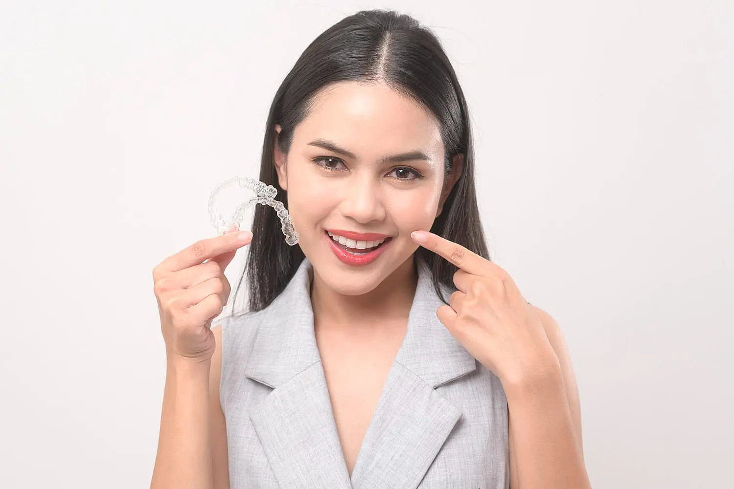 A woman smiles and points at her teeth while holding a clear Invisalign aligner in one hand, showcasing the benefits of adult orthodontics. She wears a light gray blazer in Leominster and Gardner, MA