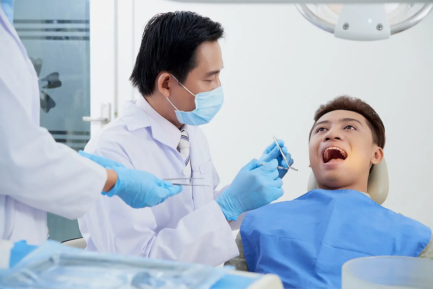 The dentist examines a patient's teeth, carefully monitoring for signs of tongue thrust, while an assistant stands nearby. Both wear masks and gloves, ensuring a sterile environment in Leominster and Gardner, MA
