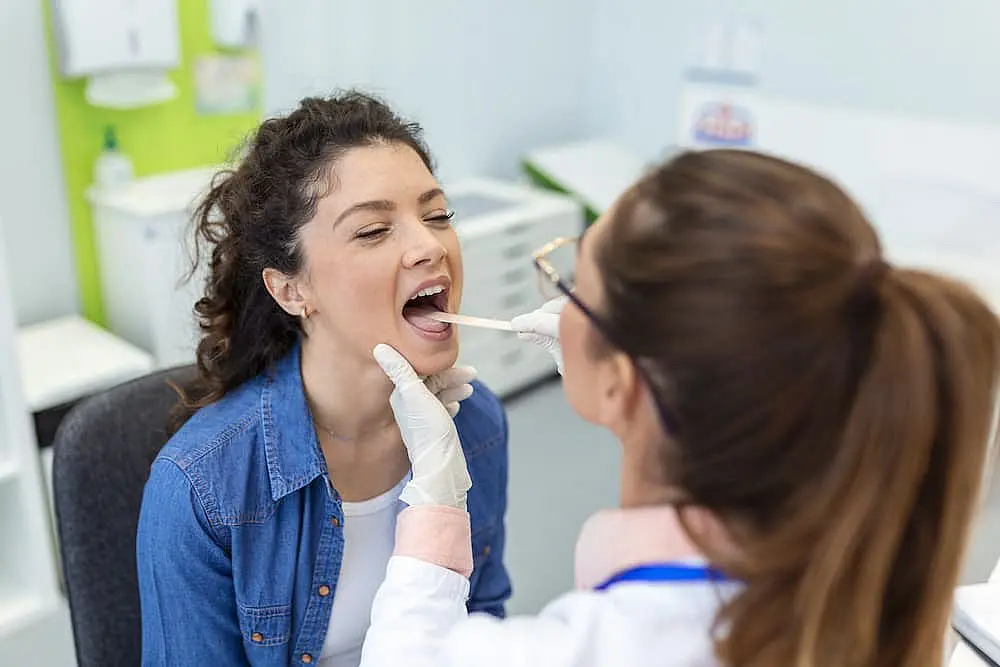 A doctor in a white coat examines a patient's throat, checking for signs of tongue thrust, using a tongue depressor in a medical office setting in Leominster and Gardner, MA