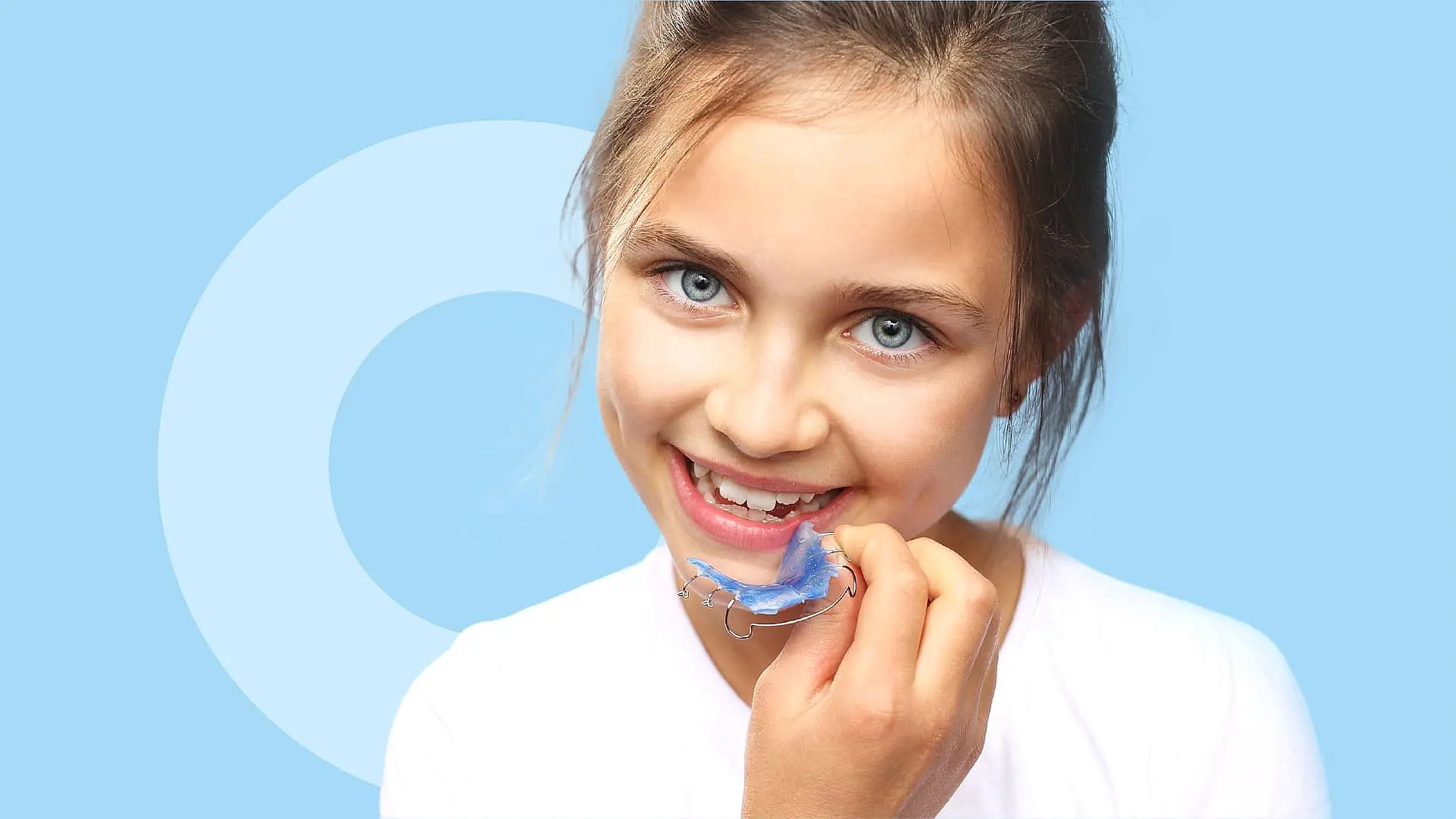 A young girl smiles, holding an orthodontic appliance—a blue dental retainer—close to her mouth against a light blue background in Leominster and Gardner, MA
