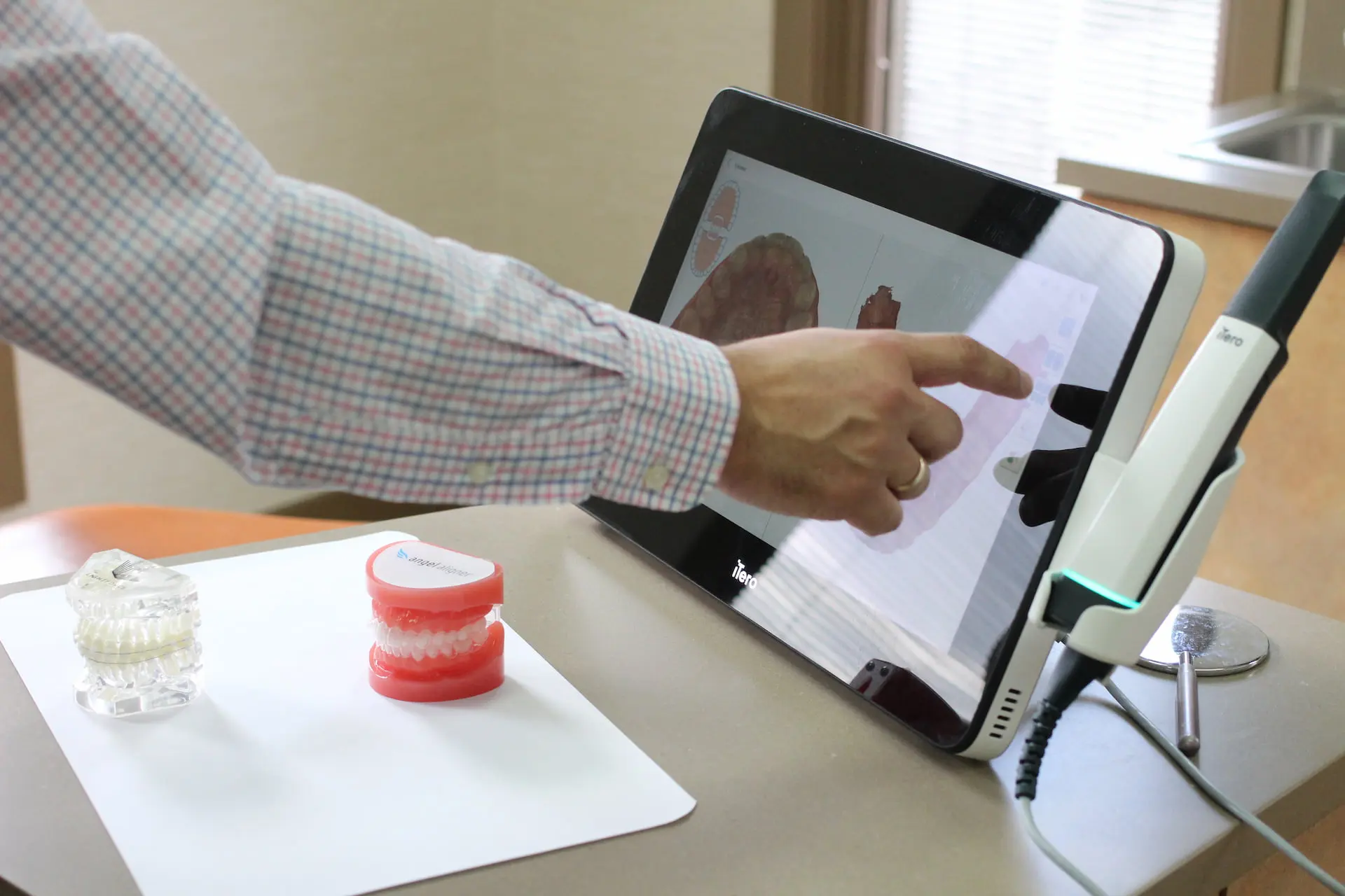 A person interacts with a touch screen displaying digital dental models, including Invisalign options, alongside two physical dental mold samples on a table in Leominster and Gardner, MA