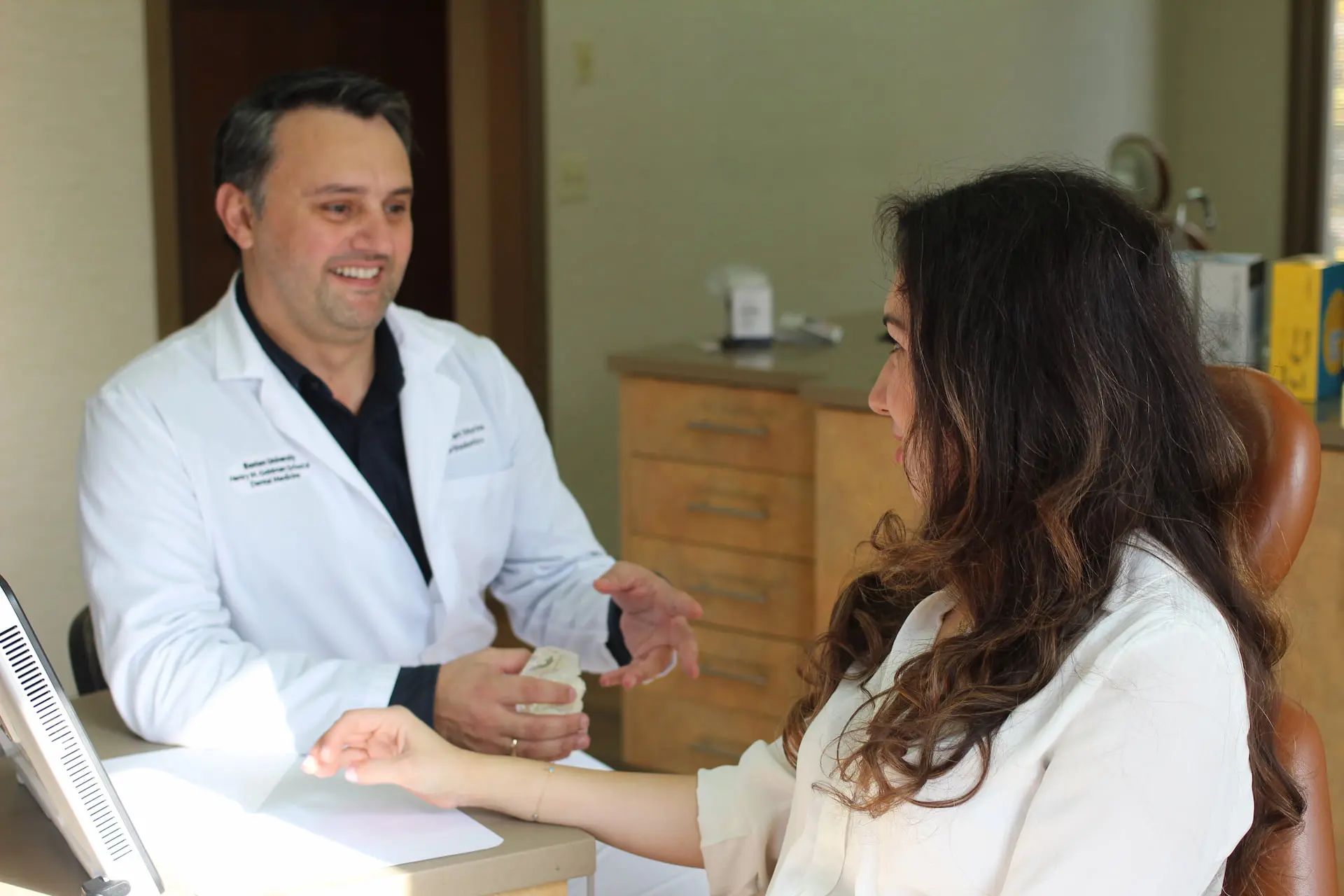 Dr. Granit Morina, in a white coat, engages with a seated woman in his medical office, gesturing warmly as he discusses her treatment options at Smile Orthodontics in Leominster and Gardner, MA