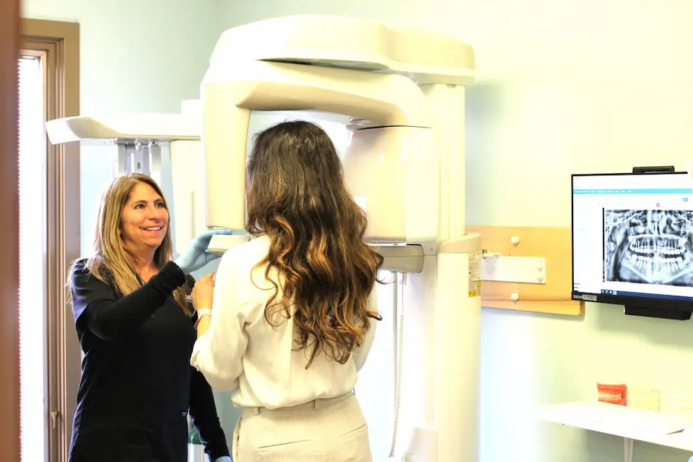 A dental technician assists a patient with a panoramic X-ray machine. A computer monitor displays a dental X-ray image at Smile Orthodontics.