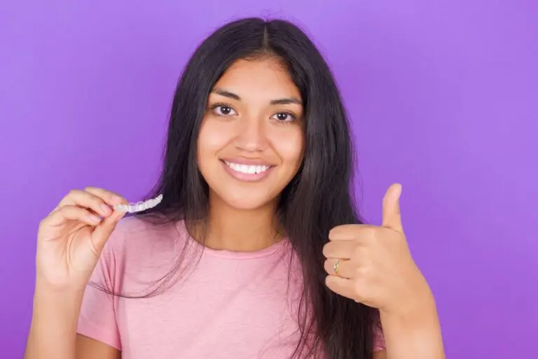 brunette girl wearing pink shirt purple background holding invisalign clear aligners - Can Invisalign Fix Overbite in Leominster and Gardner, MA.