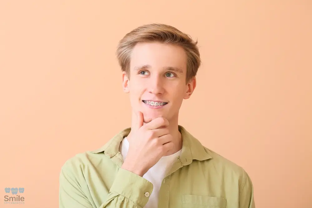 Young man with braces smiling thoughtfully, touching his chin while looking upward, standing against a peach-colored background - How to Get Braces Glue Off Teeth in Leominster and Gardner, MA