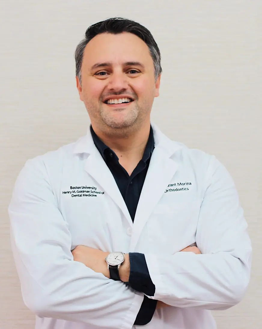 Dr. Granit Morina: Orthodontist in Gardner, MA Dr. Granit Morina, D.M.D., M.S.D stands smiling in a white lab coat, arms confidently crossed against a plain background.
