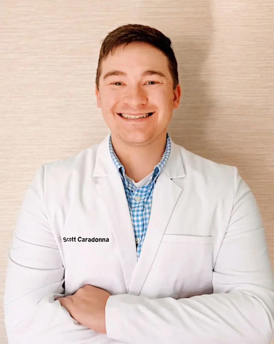 Dr. Scott Caradonna: Orthodontist in Gardner, MA Dr. Scott Caradonna stands with a smile, wearing a white lab coat over a blue checkered shirt, arms confidently crossed against a light-colored wall.